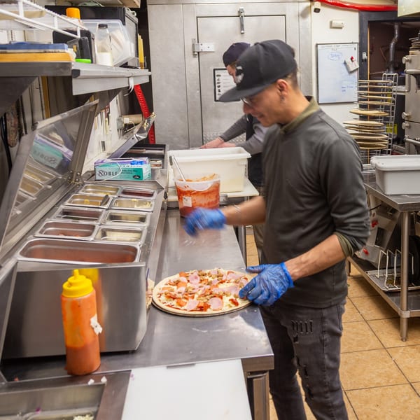 A chef in blue gloves prepares a pizza with toppings in a busy kitchen at Angel Fired Pizza.