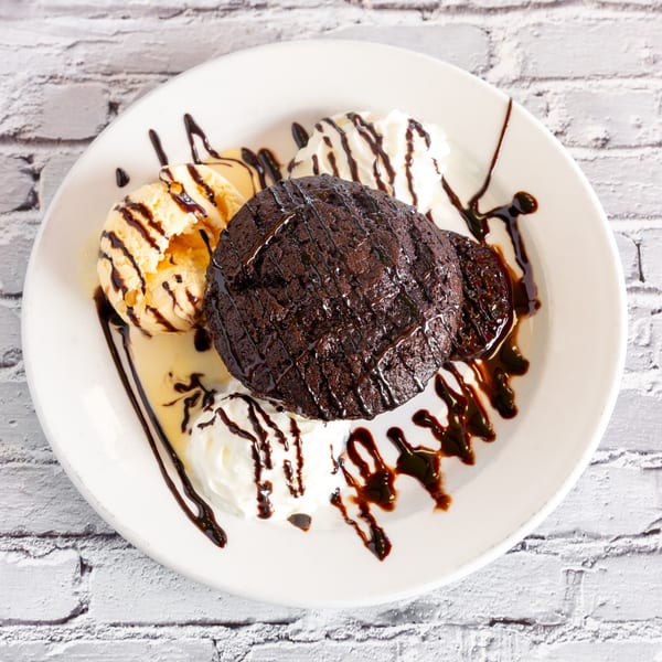 Warm chocolate brownie with vanilla ice cream, whipped cream, and chocolate syrup on a white plate.
