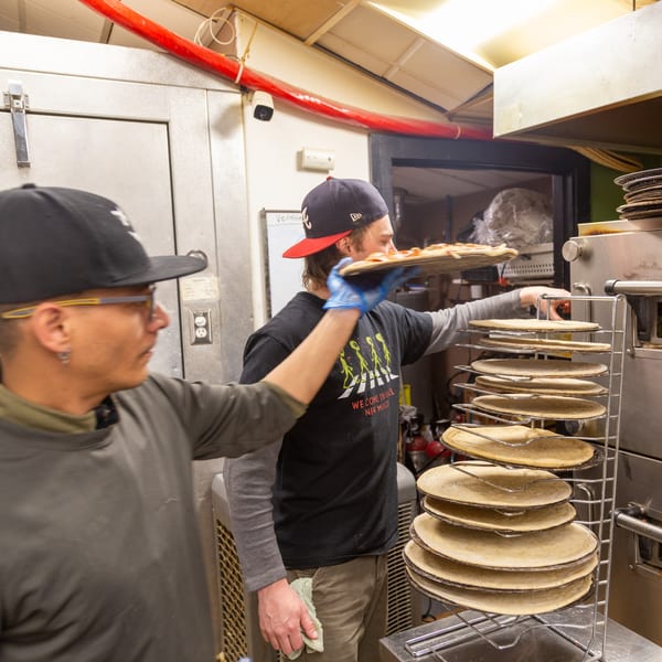 Two Angel Fired Pizza chefs preparing pizzas in a busy kitchen with pizza crusts on racks.