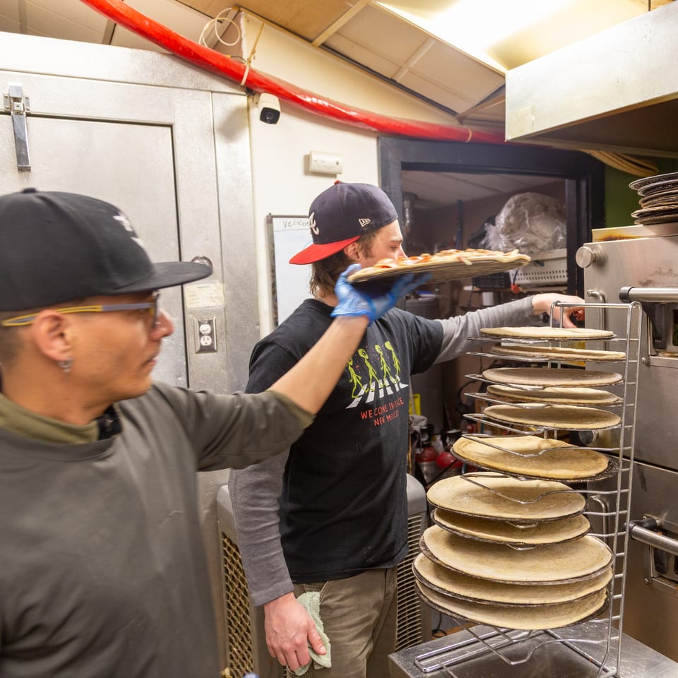 Two Angel Fired Pizza chefs preparing pizzas in a busy kitchen with pizza crusts on racks.