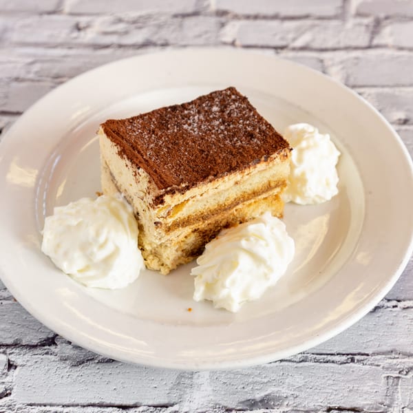 A slice of tiramisu with cocoa powder and three dollops of whipped cream on a white plate.