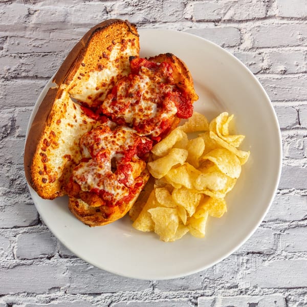 Meatball sub sandwich with melted cheese and marinara sauce, served with potato chips on a white plate.